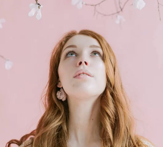 A headshot of a youngish woman with long ginger hair, looking upwards towards floral decorations.